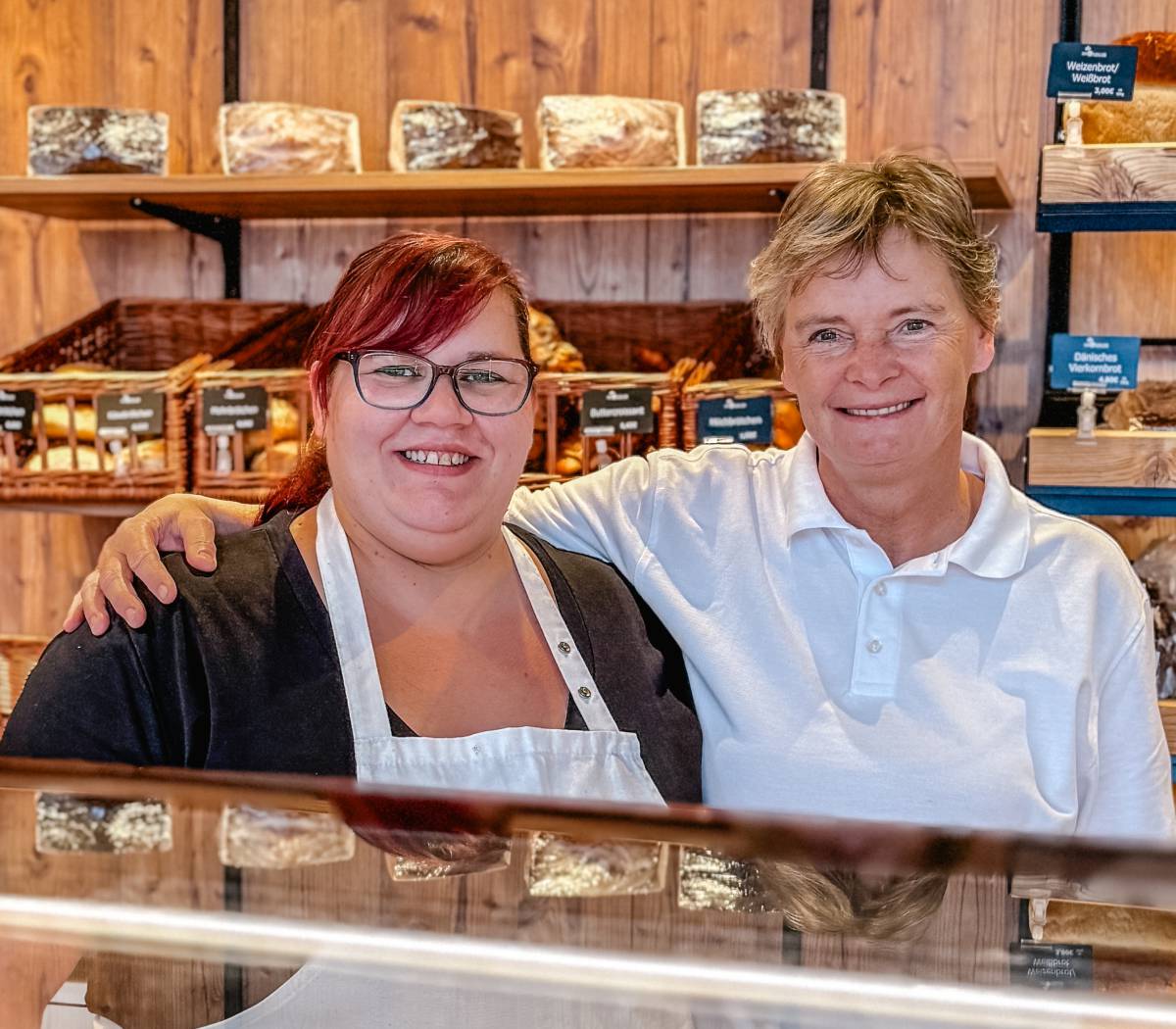Zwei freundliche Verkäuferinnen im Backshop des Ahlbäckers stehen lächelnd hinter der Verkaufstheke vor Regalen mit frisch gebackenem Brot