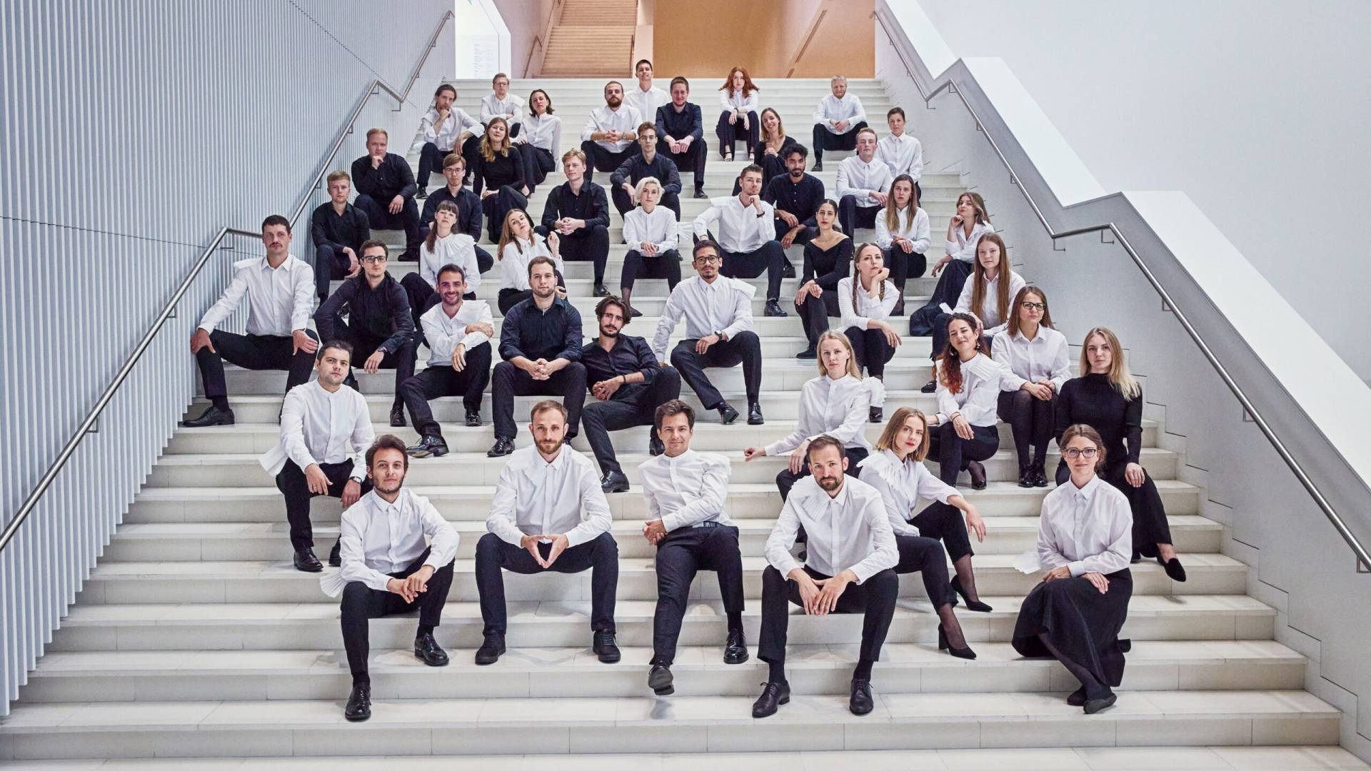 Großes Musikensemble in schwarz-weißer Kleidung sitzt auf einer Treppe in einem modernen Gebäude – Gruppenfoto im Rahmen des Usedomer Musikfestivals auf der Ostseeinsel Usedom