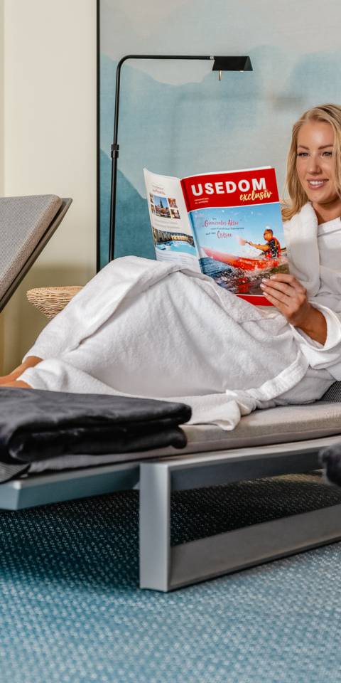 Frau im Bademantel entspannt im Ruheraum des Das Ahlbeck Hotel & Spa****s und liest eine Zeitschrift auf einer Liege