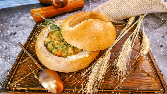 Eintopf im Brotlaib der Hotelbäckerei DER AHLBÄCKER auf einem Holzbrett, mit Ähre, Löffel und Salz- und Pfefferstreuer