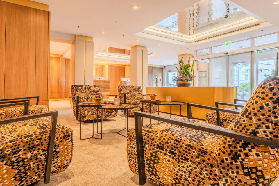 Hotellobby DAS AHLBECK HOTEL & SPA mit Schwingsesseln, Blumengesteck auf goldenem Sideboard als Dekoelement, Säulen und Rezeptionstresen im Hintergrund