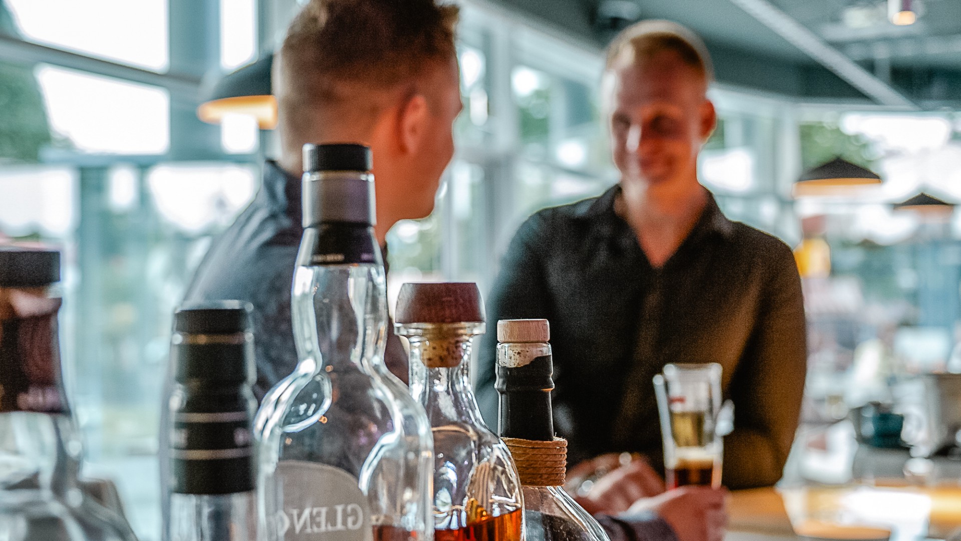 Gäste genießen Drinks an der Bar in Günter’s Bar im DAS AHLBECK HOTEL & SPA auf Usedom, mit stilvoll arrangierten Spirituosenflaschen im Vordergrund