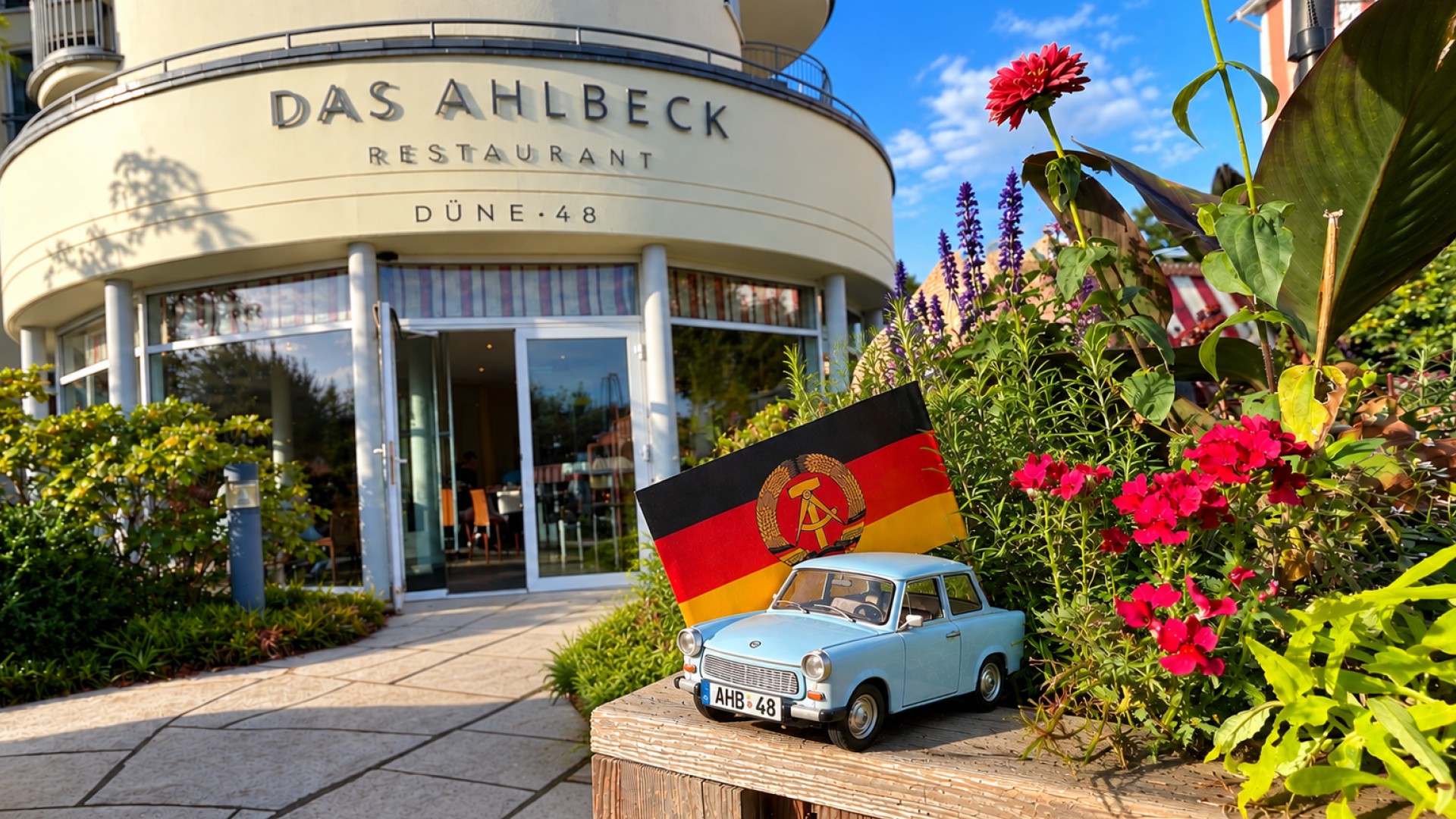 Restauranteingang DÜNE 48 mit offener Tür, davor Blumen, ein hellblauer Trabant und eine DDF Fahne