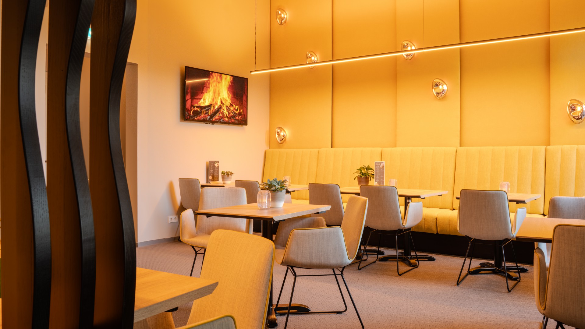Stühle und Tische mit Kerze, Blume und Speisekarte in der Café-Bar Günter`s mit gelber Sitzbank und gelb gepolsterter Rückwand im Hintergrund und Kaminfeier am an der Wand hängenden TV 